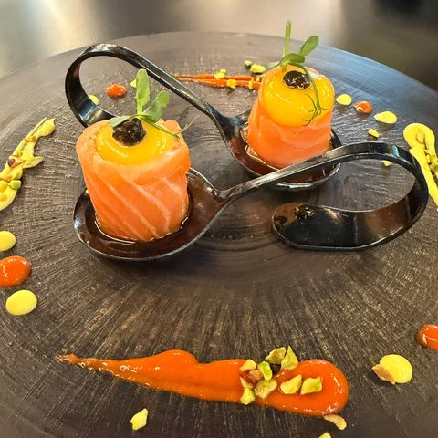 Involtini di salmone farciti con crema al mango e uova di lompo nere, serviti su cucchiai di metallo intrecciati